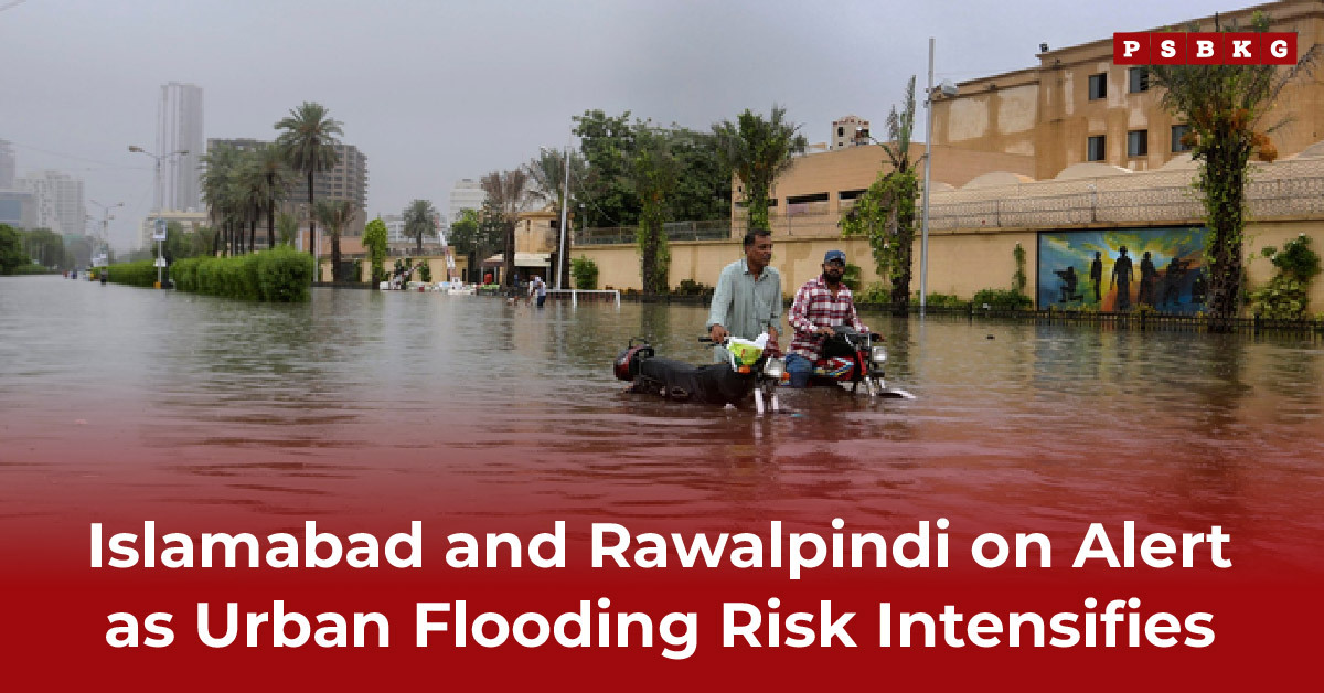 Two men navigate a flooded street on a motorcycle during urban flooding, with submerged buildings and palm trees around them under a gray, overcast sky.
