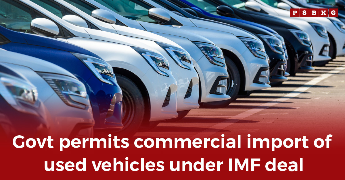 Row of parked vehicles with a banner announcing government approval for used vehicle import policy under IMF deal.