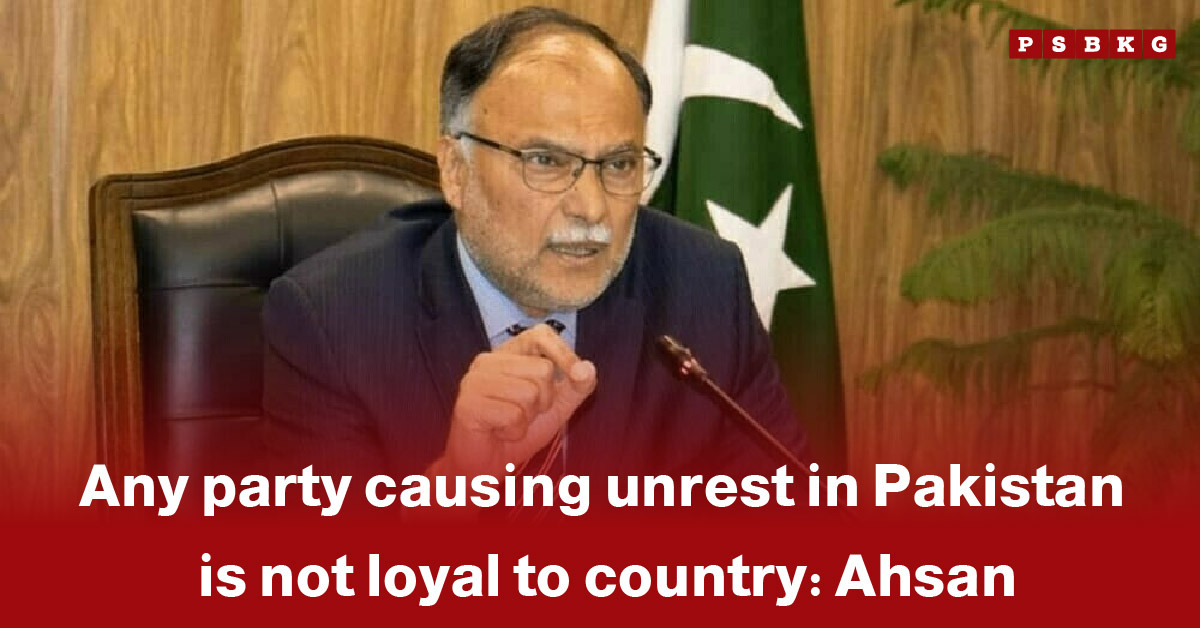 A person seated at a desk, gesturing while addressing concerns over national unrest in Pakistan, as Ahsan warns against unrest, with wooden paneling and a green-and-white flag in the background.